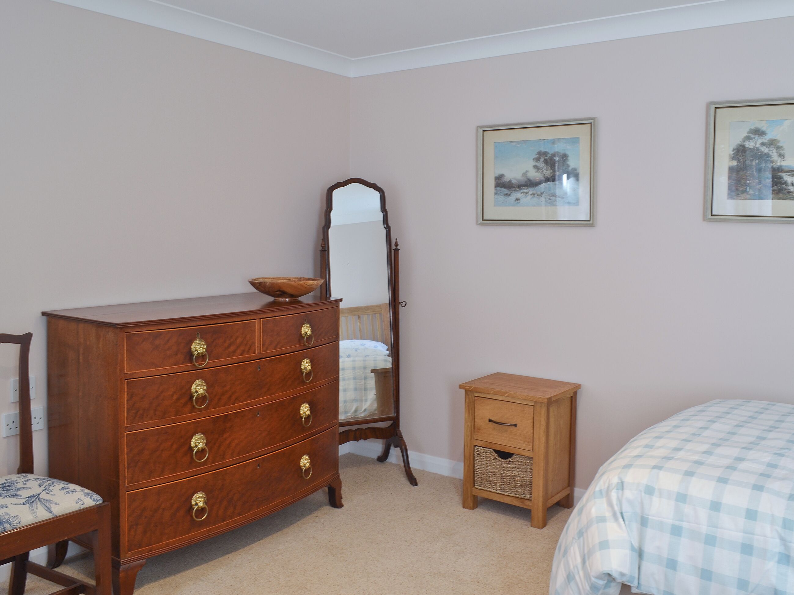 A bedroom with a wooden dresser, a mirror, a small bedside table, and two framed pictures on the wall.