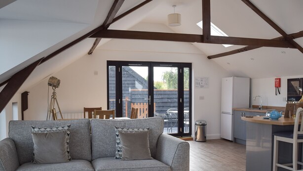 A loft-style living space with a grey sofa, a patterned ottoman, a dining area with chairs, a kitchen with a sink and a blue kettle, and a wooden ceiling structure. - 2 bedroom accommodation in Beccles, near Gillingham (Beccles, near Gillingham)