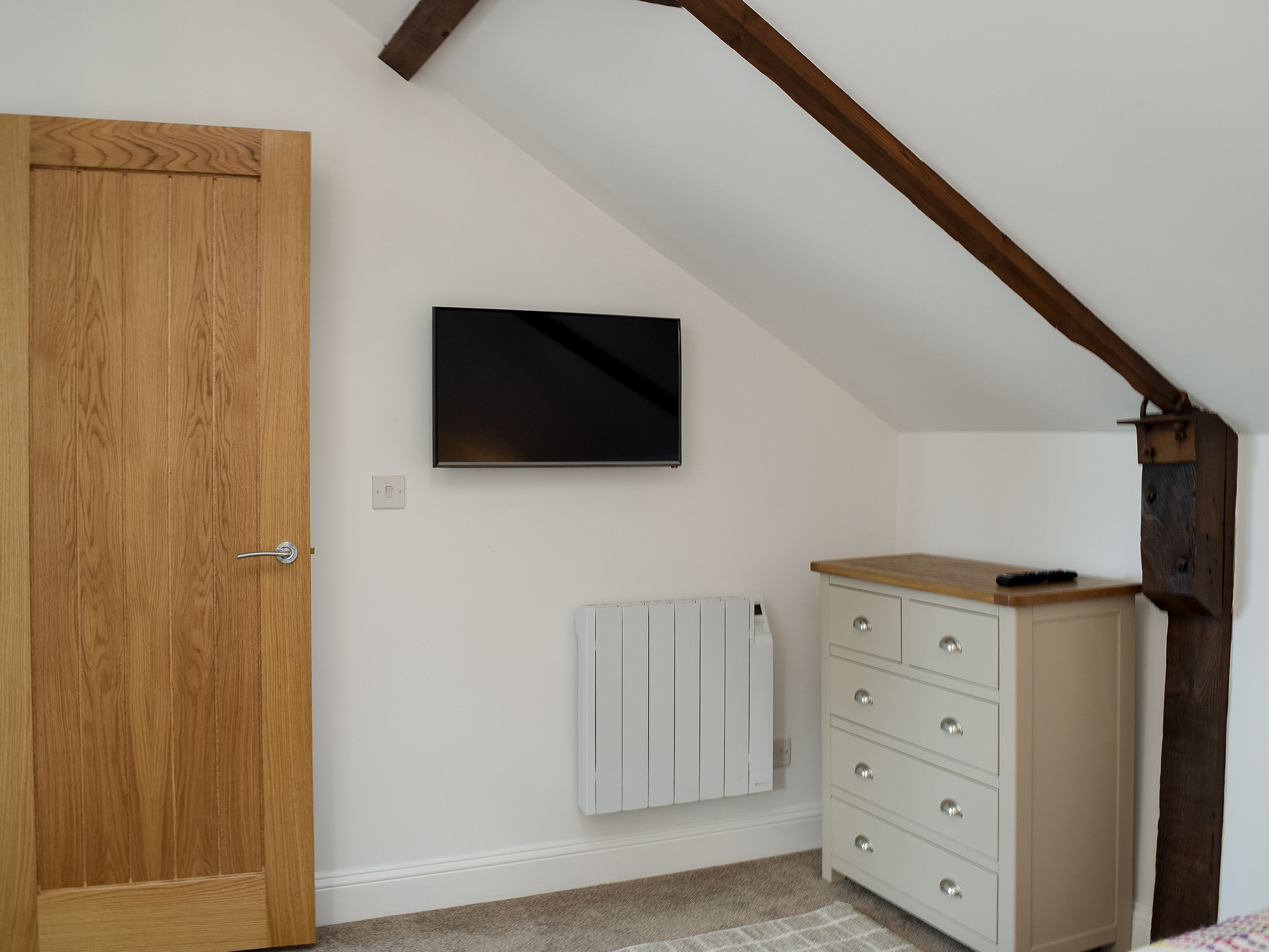 Quarto com porta de madeira, radiador branco, cómoda branca e televisão fixada na parede.