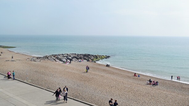 Ein Kiesstrand mit Menschen, die darauf laufen und sitzen, einem Felssteg und ruhiger See.
