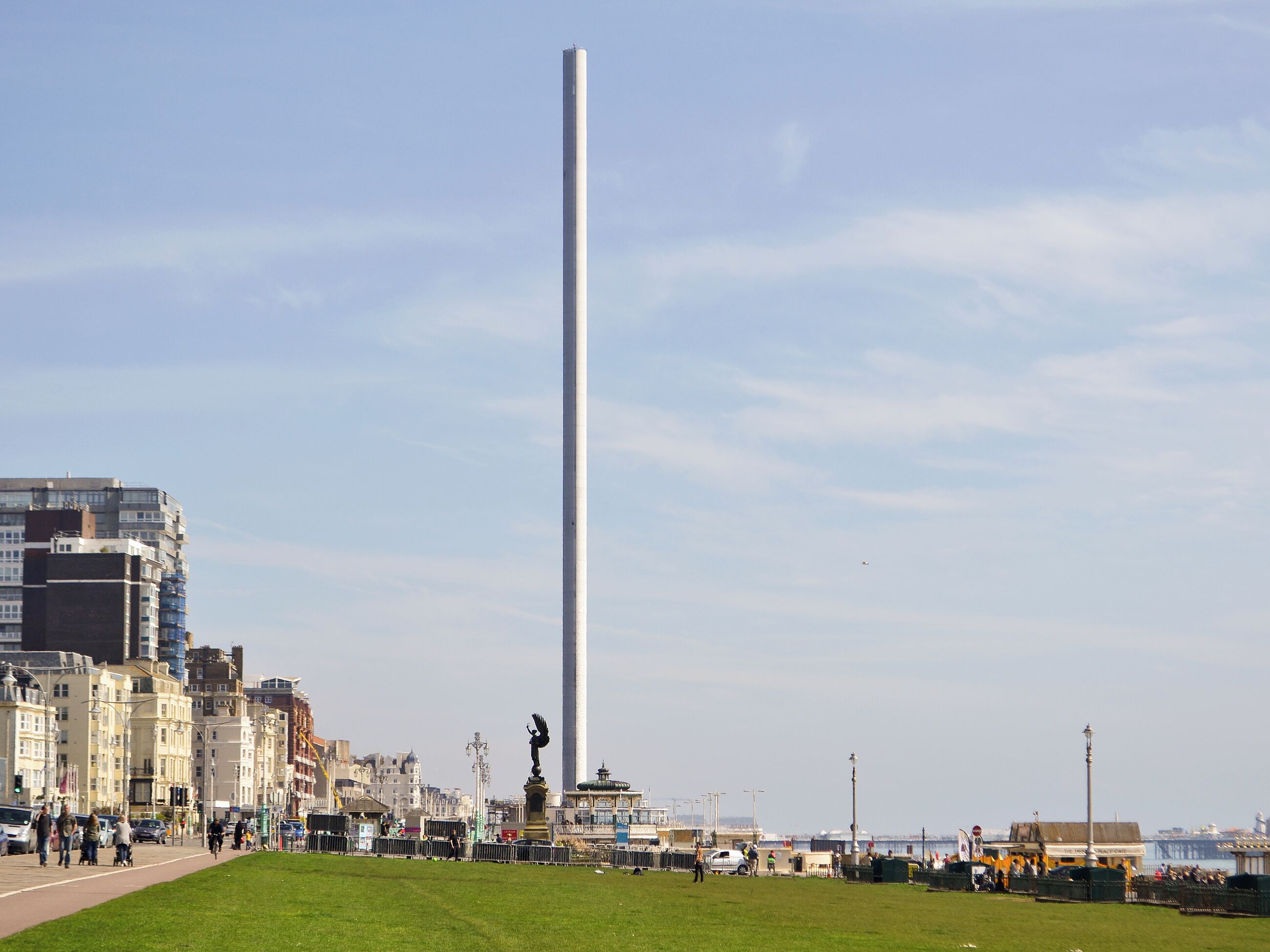 A tall monument in a public square with buildings and a body of water in the background.