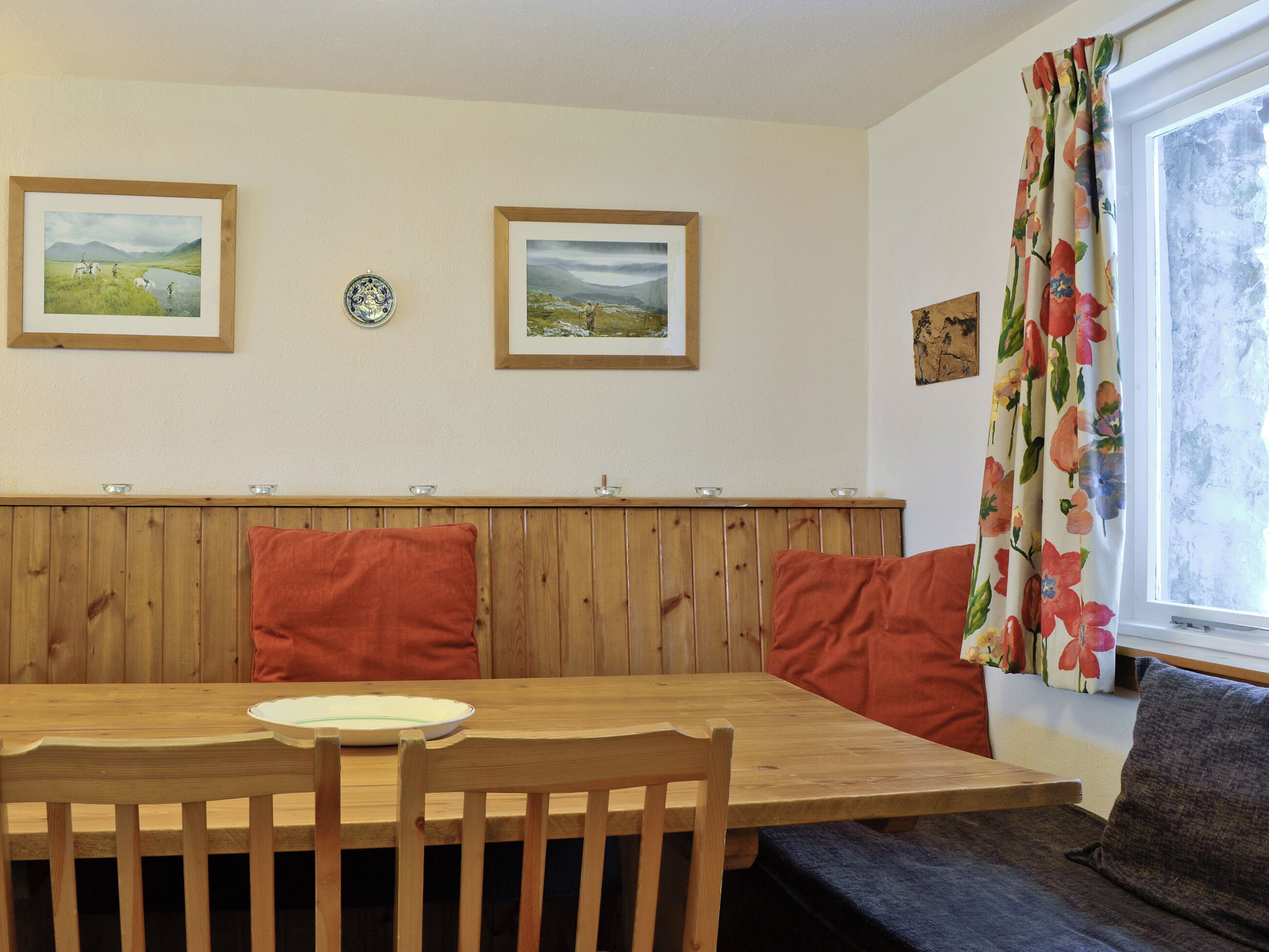 Un coin repas comprenant une table en bois, deux chaises, un banc avec des coussins et une fenêtre agrémentée de rideaux à motifs floraux.
