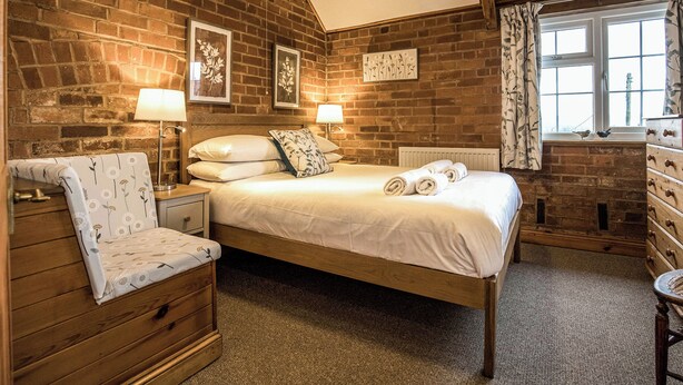 A bedroom with a brick wall, a wooden bed, a nightstand with a lamp, a wooden chair, and a dresser with a mirror. - 2 bedroom accommodation in Ledbury (Ledbury)