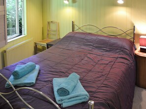 Una habitación con una cama, dos toallas enrolladas, una silla, una mesa y una ventana.