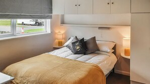 Een net opgemaakt bed met een beige deken en kussens, twee nachtlampjes en een raam met uitzicht op een geparkeerde vrachtwagen erbuiten.
