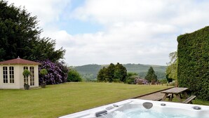 A hot tub in a garden with a gazebo and chairs, surrounded by trees and hills.