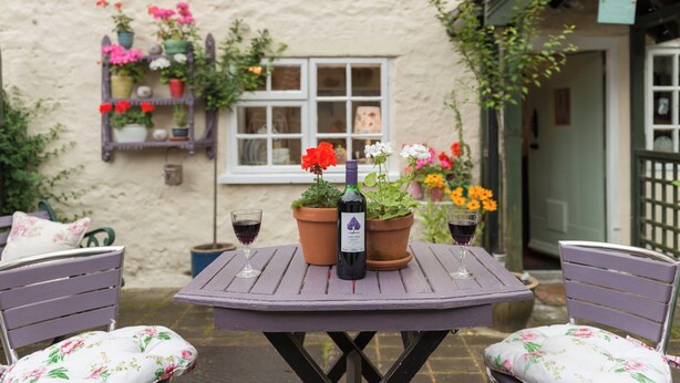 A small outdoor table with a bottle of wine and glasses, surrounded by chairs with floral cushions, set against a house with a white window and flower boxes. - Brewery House Cottage (Bishop Middleham, near Durham)
