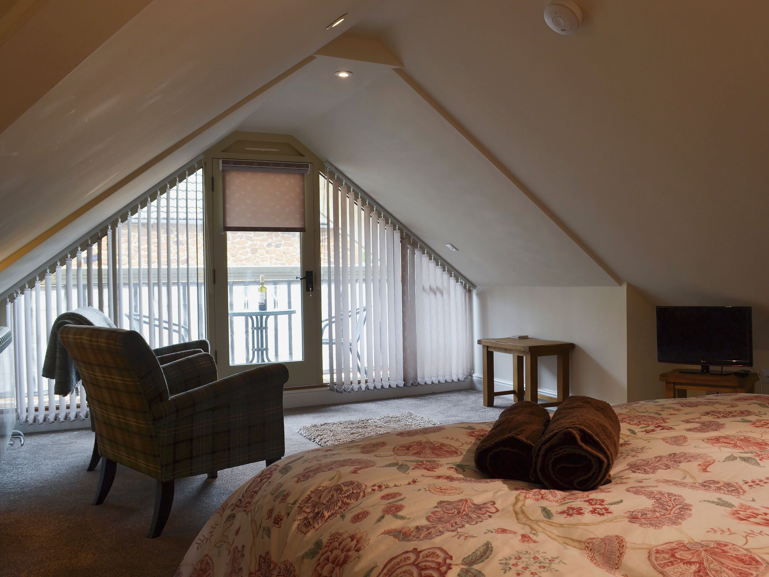 A bedroom with a sloped ceiling, a bed with floral bedding, a plaid chair, a small table, and a television.