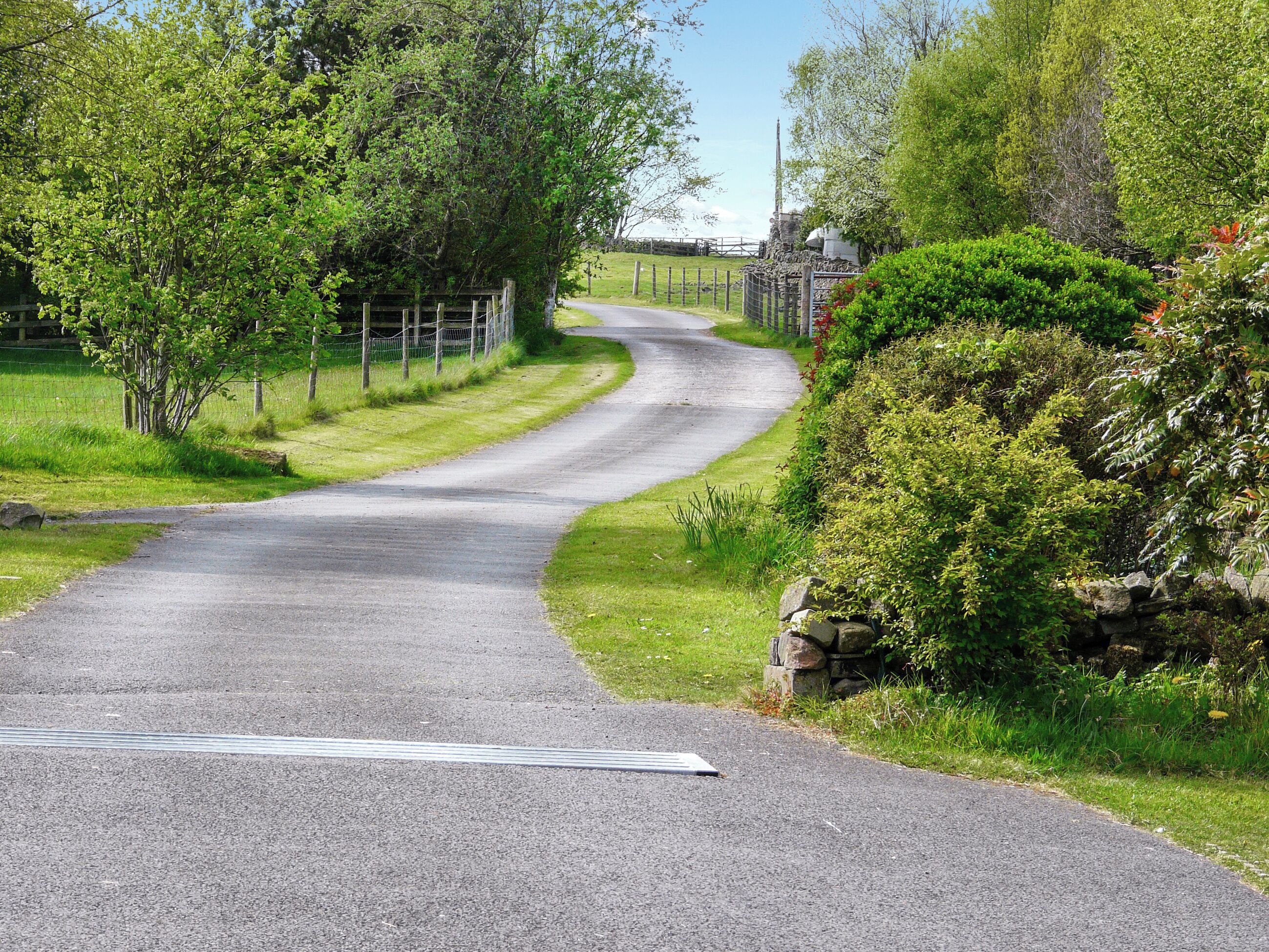 A rural road curving through a green landscape with a fence on one side and a stone wall on the other.