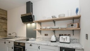 Una cocina moderna con armarios blancos, horno empotrado, estufa, cafetera y tostadora. Hay repisas de madera sobre la encimera con vajilla y vasos.