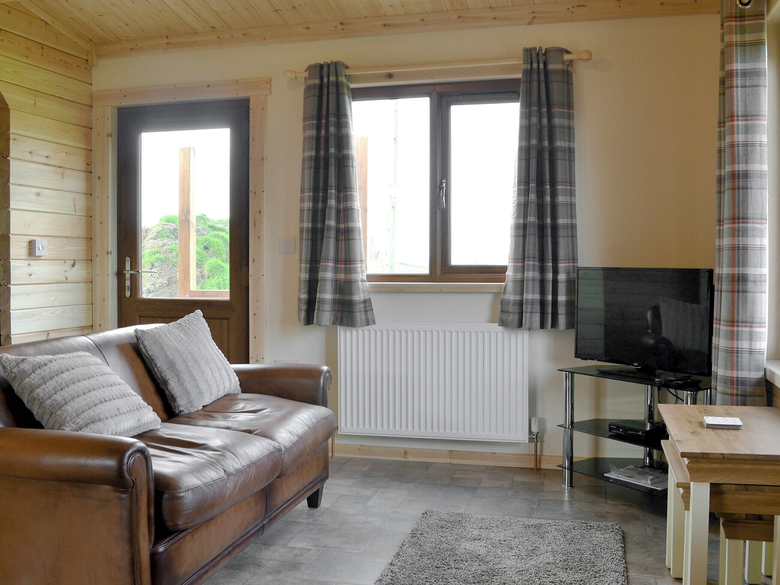 A cozy room with a brown leather sofa, a television on a stand, and a window with plaid curtains.