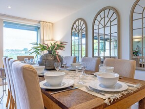 Un espace repas avec une grande table dressée pour six personnes, entourée de chaises confortables, d’un vase rempli de fleurs et de grandes fenêtres en arc de cercle.
