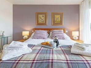 Un lit bien fait, recouvert d’une couverture à carreaux, avec deux oreillers, un plateau contenant un croissant et une tasse de café, et deux lampes de chevet.