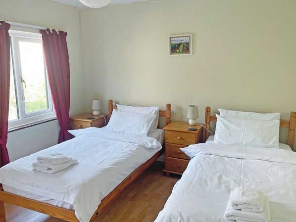 A room with two beds, wooden headboards, bedside tables with lamps, a small painting on the wall, and a window with curtains.