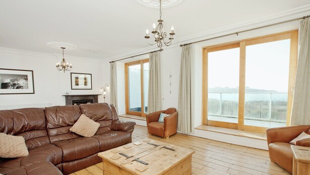 Een ruime woonkamer met een bruine leren bank, een houten salontafel en een groot raam met uitzicht op zee.