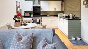 Et moderne køkken med hvide skabe, en sort komfur og en mikroovn. Der er et spisebord med stole og en grå sofa med mønstrede hynder.
