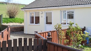 A single-story house with a tiled roof, a front door, and a small garden area with plants and a wooden fence. - 2 bedroom accommodation in Chivenor Cross, near Barnstaple (Chivenor Cross, near Barnstaple)