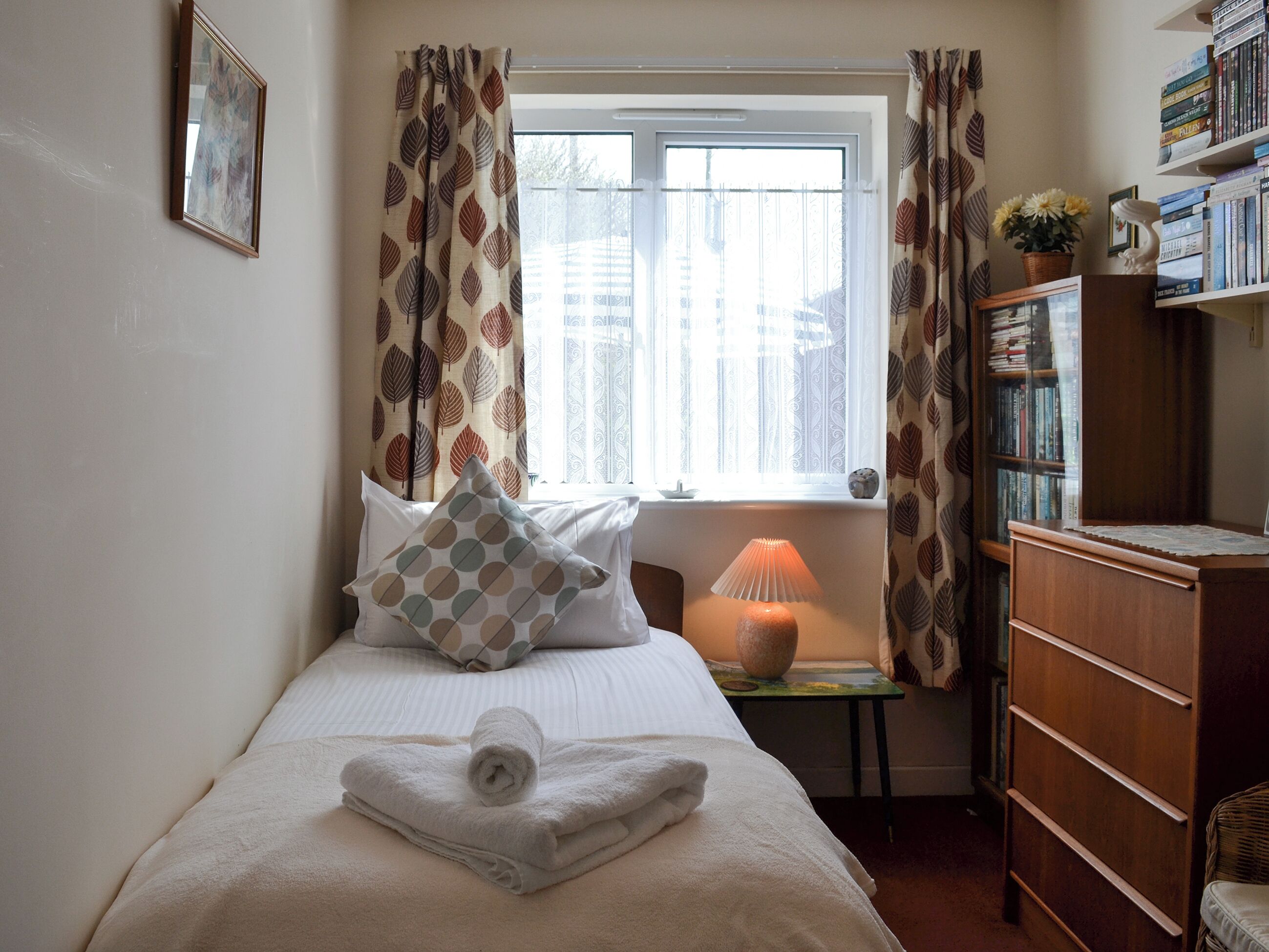 A neatly made bed with a rolled towel, a bedside lamp, and a bookshelf with books and decorative items.