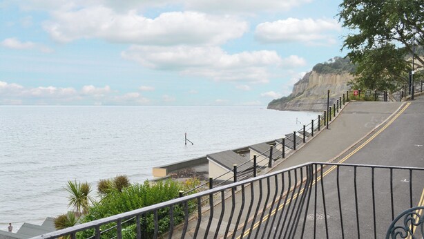 Une promenade en bord de mer, agrémentée d’une rambarde en métal, d’une table et de chaises, d’où l’on peut admirer la mer et les falaises.