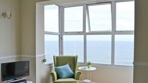 Une pièce avec une grande fenêtre, un fauteuil vert, une petite table ronde et un téléviseur posé sur un meuble blanc.