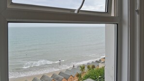 Une vue sur la plage, prise à travers une fenêtre, qui inclut la mer, les cabanes de plage et une personne se promenant le long du rivage.
