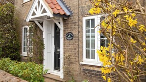 Uma casa de tijolos com porta branca, um pequeno alpendre e uma placa com a inscrição 'HAPENY COTTAGE'.