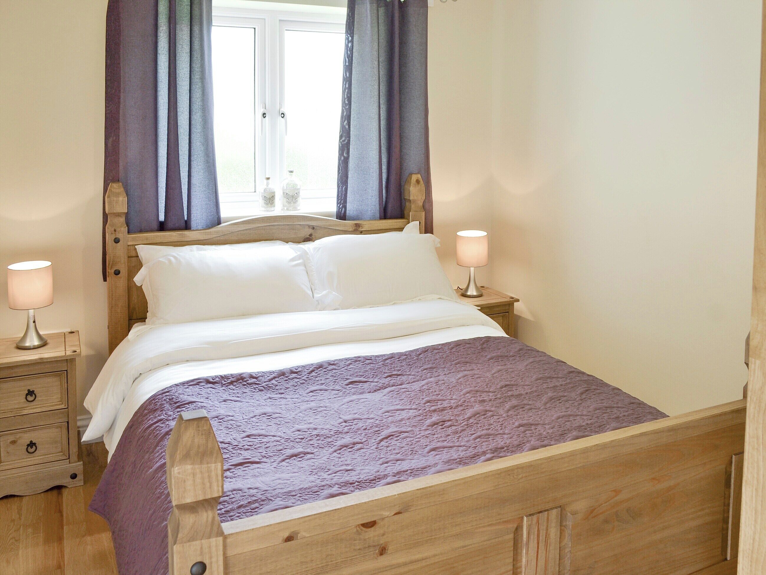 A wooden bed with white bedding and a purple blanket, flanked by nightstands with lamps, in a room with a window and curtains.