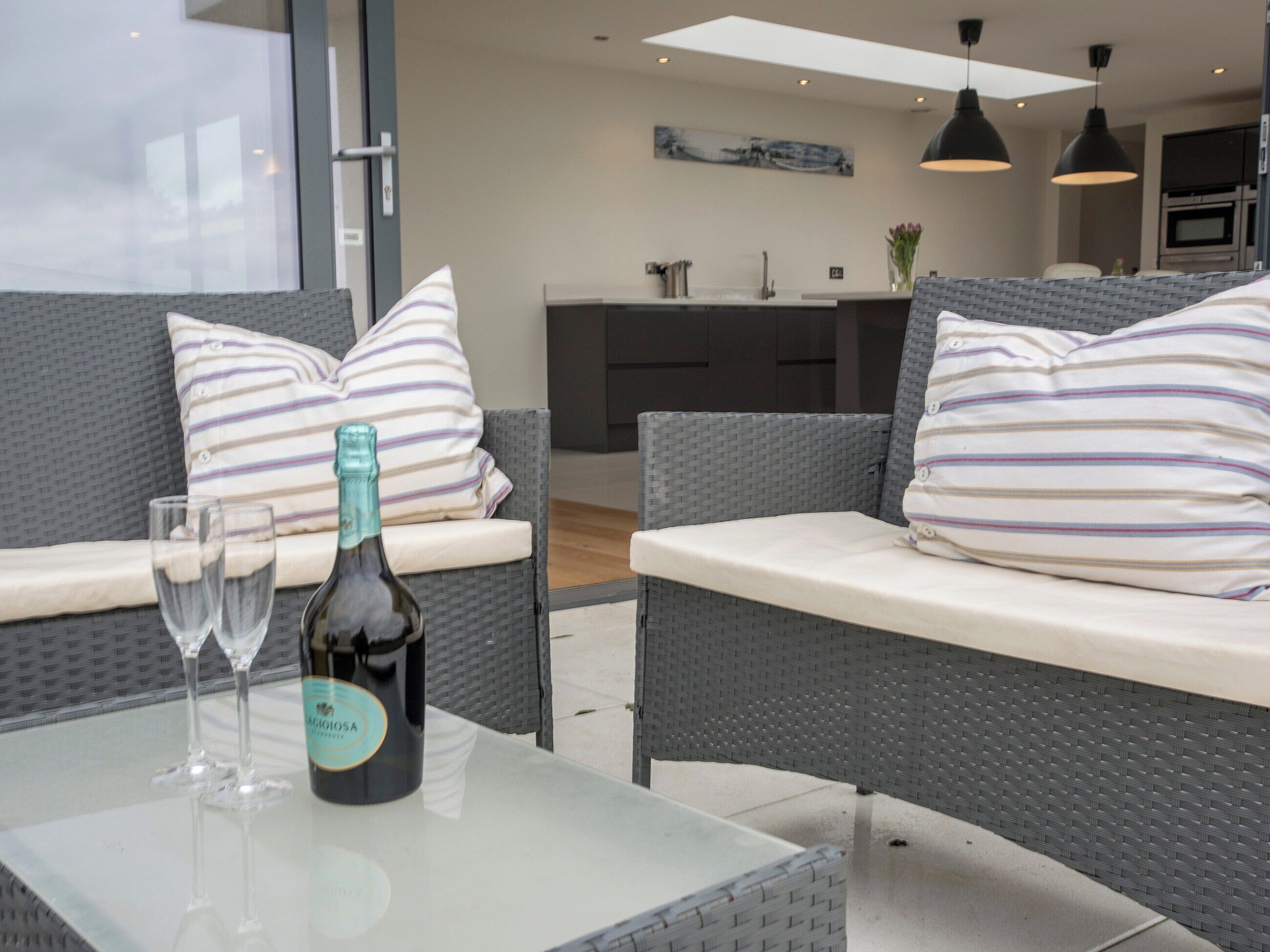 Un espace extérieur moderne aménagé avec des meubles en osier, une table en verre et une bouteille de vin.