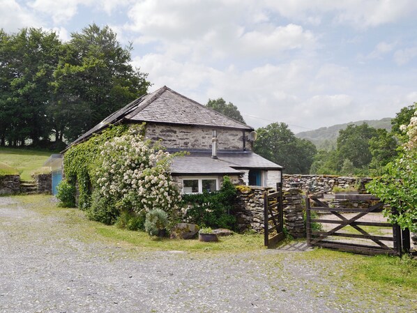 A stone house with a sloped roof, surrounded by greenery and a gravel path.