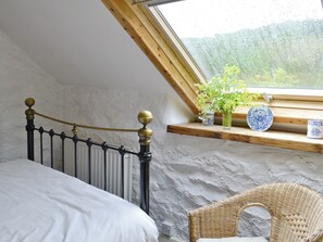 A bedroom with a bed, a wicker chair, a window with a view of greenery, and a shelf with decorative items. - 2 bedroom accommodation in Betws-y-Coed (Betws-y-Coed)