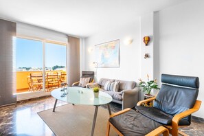 A modern living room with a glass table, leather armchair, sofa, and a large window offering a view of the city.