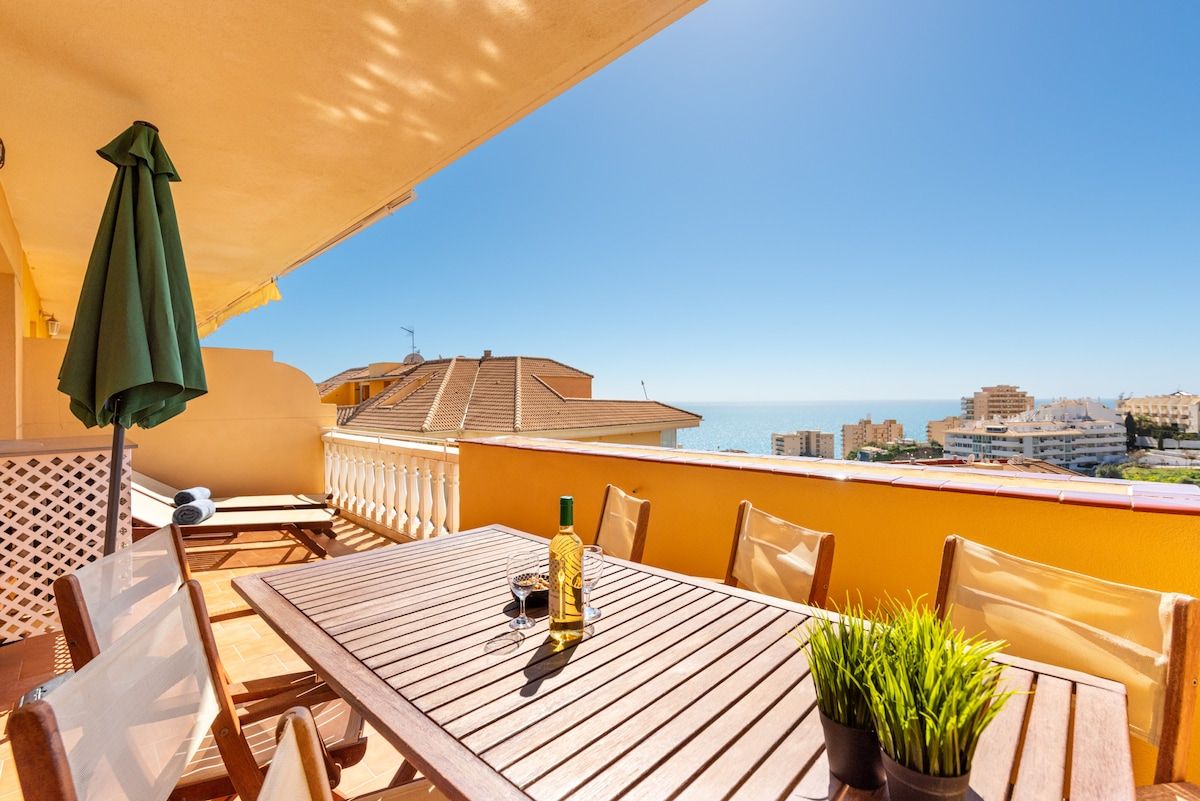 Une table en bois, des chaises et un parasol vert sur un balcon donnant sur un paysage urbain.