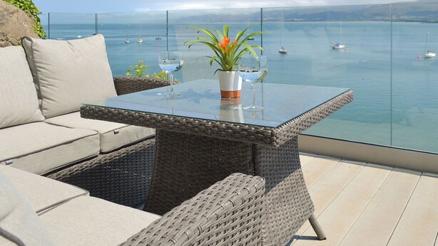 A glass-top table with two potted plants, a wicker sofa, and a view of the sea with boats.