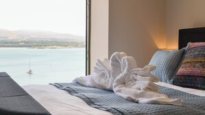 A hotel room with a bed, a view of a body of water, and a sailboat in the distance.