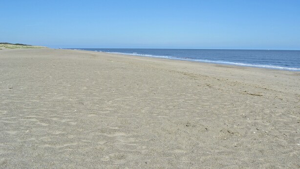 Ein weiter, sandiger Strand mit sanften Wellen und wolkenlosem, blauem Himmel.