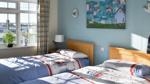 Ein Schlafzimmer mit zwei Betten, einem Fenster mit Vorhängen, einem Gemälde mit Küstenmotiv und einer Lampe auf einem Nachttisch.