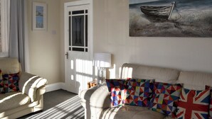 Ein Wohnzimmer mit einem beigen Sofa, einem gemusterten Kissen, einem Bild eines Bootes am Strand und einer Tür mit einer satinierten Glasscheibe.