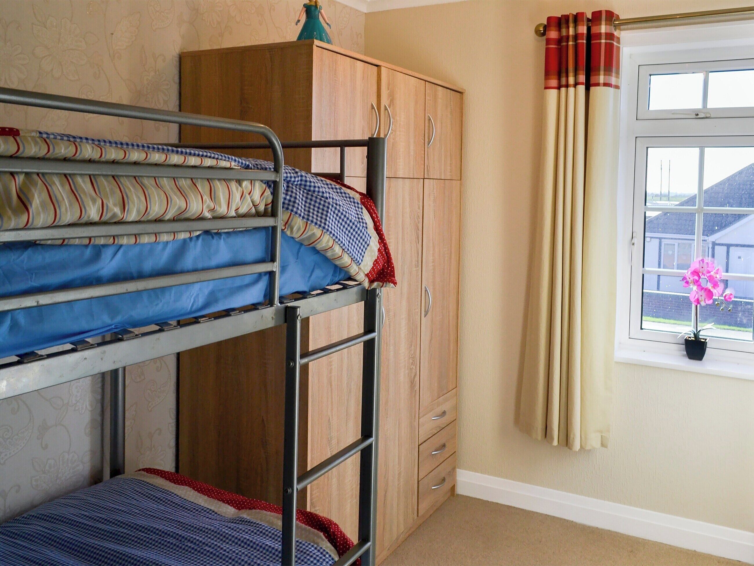 Ein Stockbett mit gemusterten Betttextilien, ein Holzschrank, ein Fenster mit Vorhängen und ein Blumentopf.