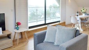 Una sala moderna con un sofá gris, una mesita blanca con un jarrón de flores, y un comedor con mesa y sillas.