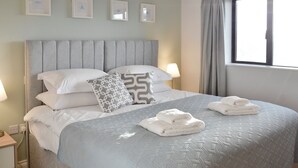 Una cama bien hecha con cabecera gris, almohadas blancas y una manta estampada. Hay dos lámparas de noche, una ventana con cortinas y una mesita de noche pequeña con una lámpara.