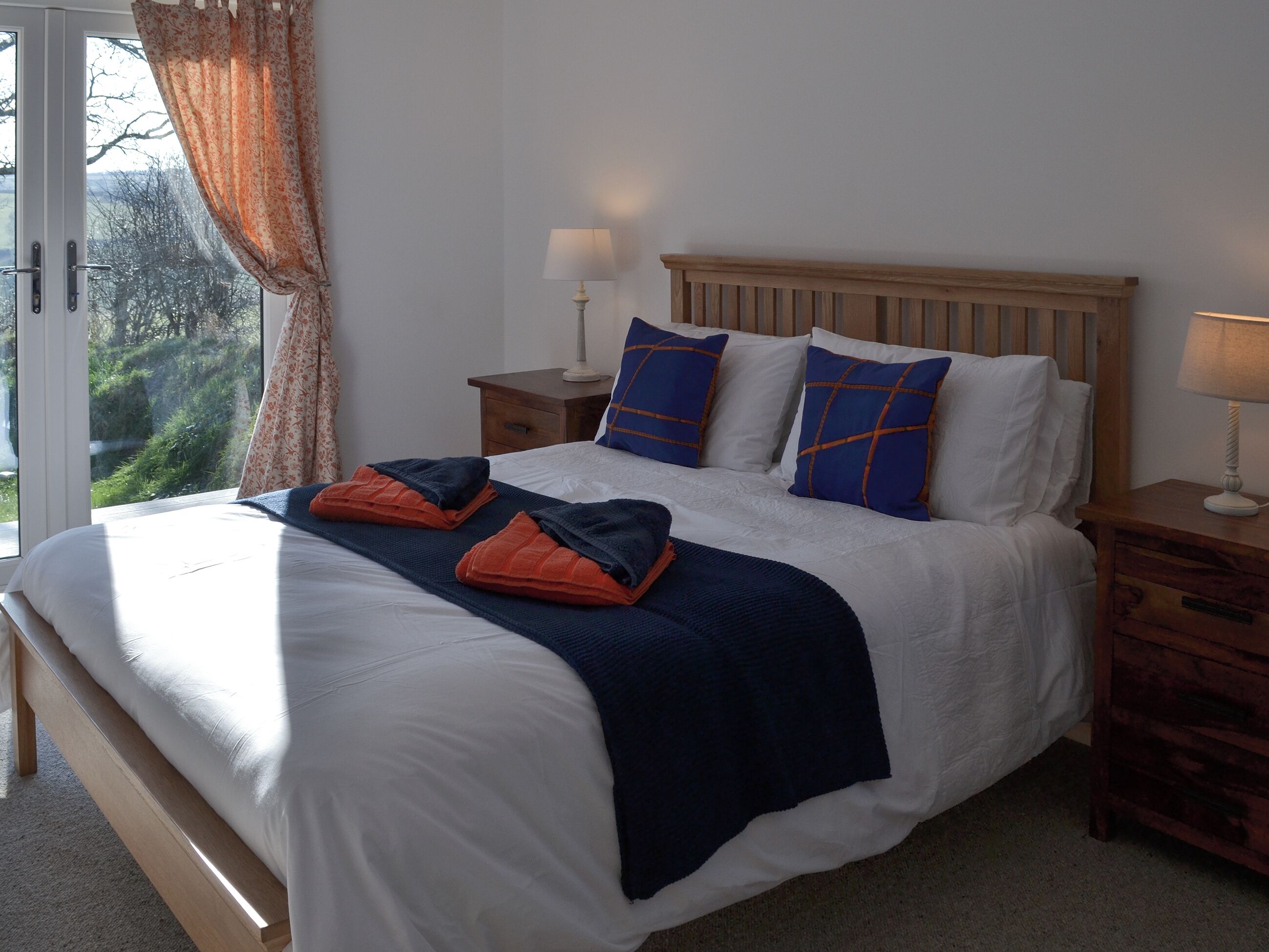 A neatly made bed with a wooden headboard, two bedside tables with lamps, and a view of greenery outside.