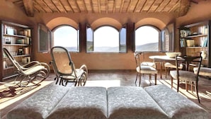 A room with a high ceiling, arched windows, a sofa, a chair, a table, and bookshelves. - VILLA CHIANTIVERTINE (Vertine)
