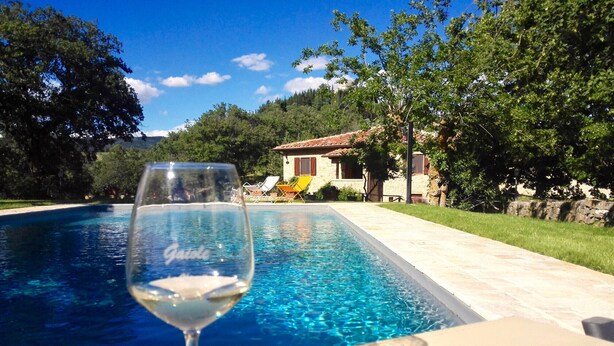 A clear swimming pool with a glass of white wine on a table, surrounded by greenery and a stone building in the background. - VILLA CHIANTIVERTINE (Vertine)