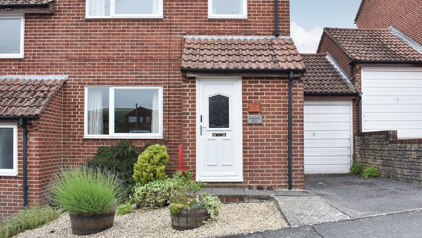 A two-story brick house with a white front door, a garage door, and a small garden with potted plants. - 3 bedroom accommodation in Preston, near Weymouth (Preston, near Weymouth)