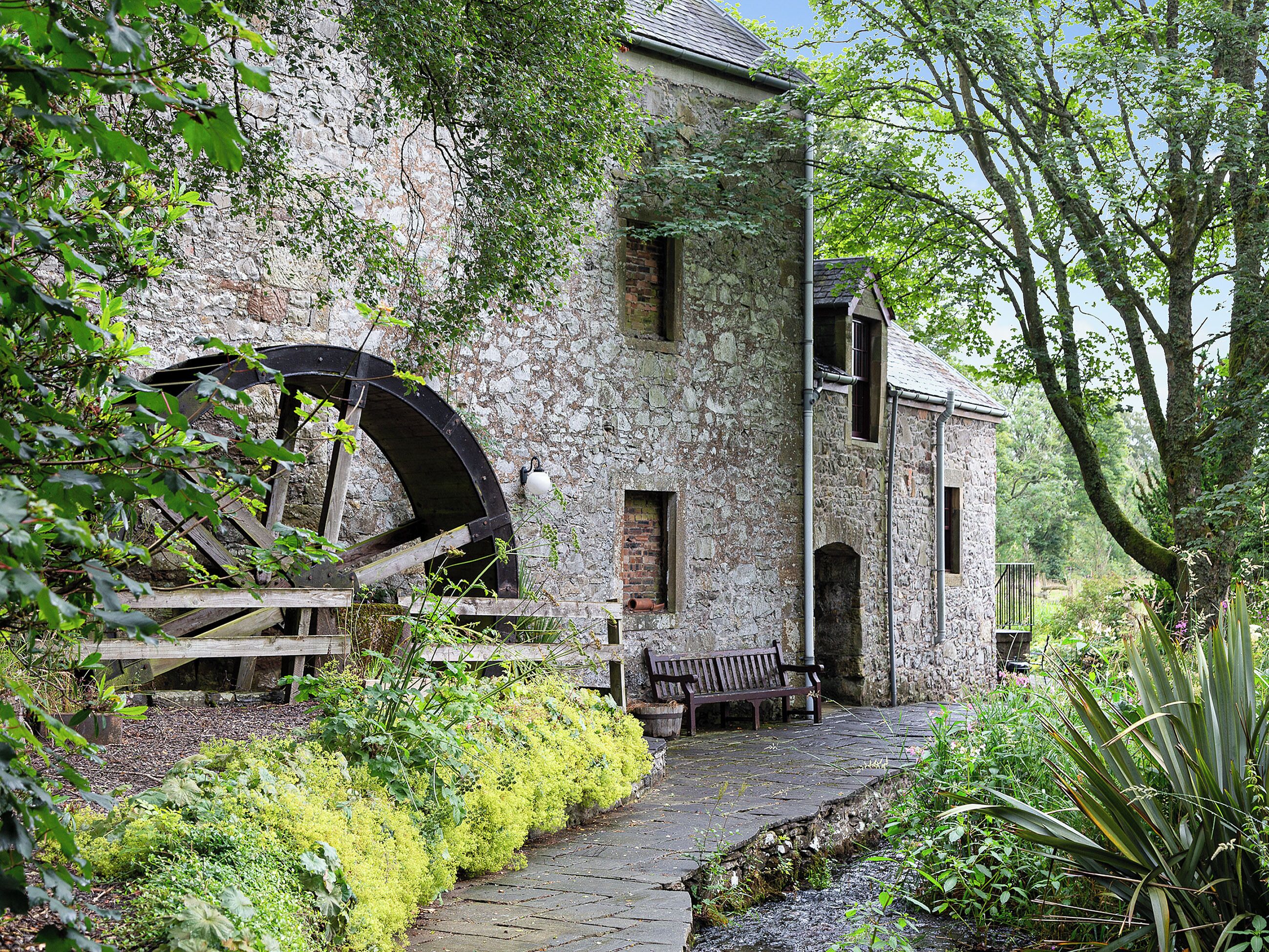 A stone building with a large water wheel, a bench, and a pathway surrounded by greenery.