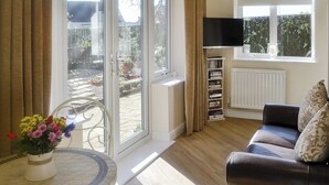 Ein Wohnzimmer mit einer Couch, einem an der Wand befestigten Fernseher, einem Bücherregal und Blick ins Freie durch eine Schiebetür.