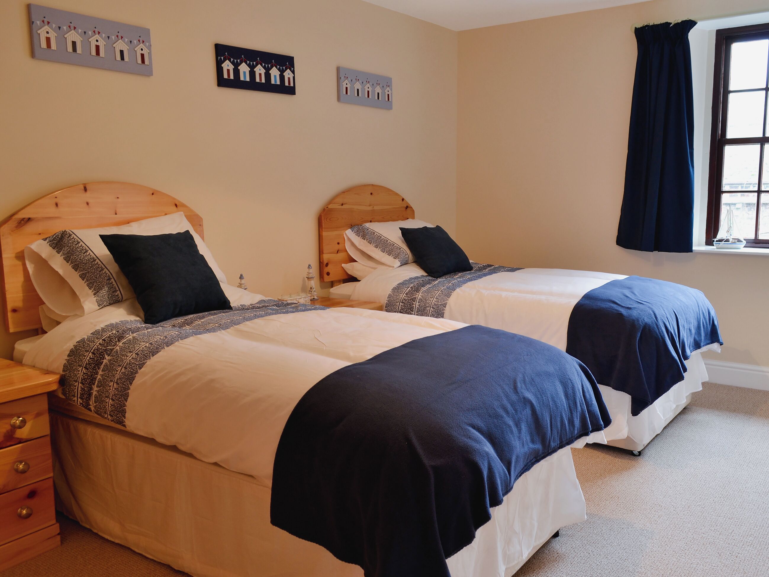 Une chambre double avec deux lits en bois, chacun doté d’une tête de lit, de draps blancs et de couvertures bleu foncé. On y trouve deux tables de chevet avec des lampes, une commode en bois et une fenêtre avec des rideaux foncés.