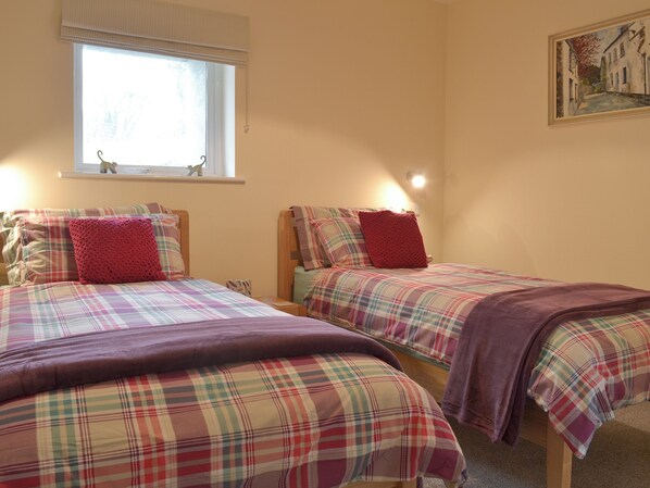 A bedroom with two beds, each with a plaid bedspread and a red pillow. There is a window with a view of trees and a small figurine on the sill.