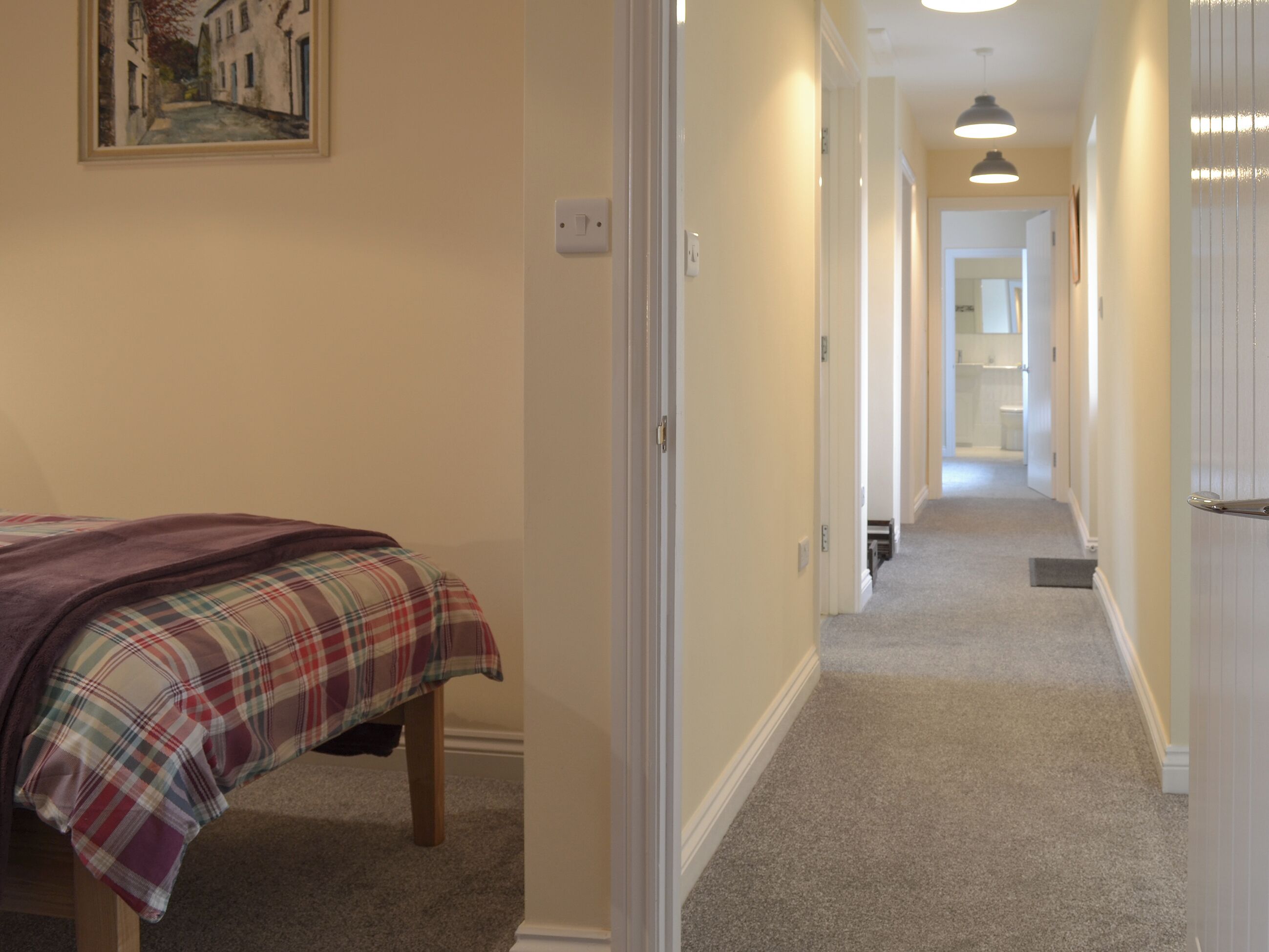 A narrow hallway with carpeted floors, a bed with a plaid bedspread, and a door on the right.