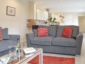 A modern living room with a grey sofa, red cushions, a glass coffee table with a wine bottle and glasses, and a kitchen in the background.